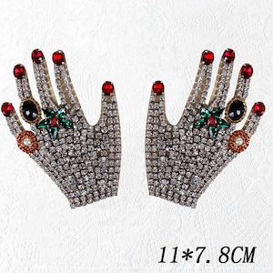 以下が含まれることがあります： 赤いマニキュア、緑と黒の宝石、真珠のアクセントが付いた、ラインストーンで覆われた2つの手形のアップリケ。アップリケは11 x 7.8センチメートルです。