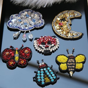 May include: Six colorful beaded iron-on patches, including a cloud with raindrops, a crescent moon, a ladybug, a butterfly, a bee, and a red panda.