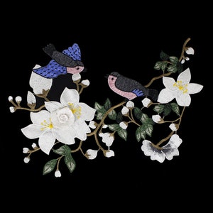 May include: Embroidered patch featuring two birds perched on a branch with white flowers. The birds are blue, pink, and black. The flowers are white with yellow centers.
