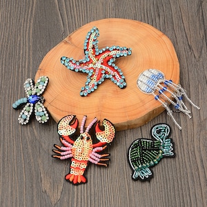 May include: A collection of beaded and sequined sea creature patches, including a red starfish with blue accents, a lobster, a jellyfish, a dragonfly, and a sea turtle. The patches are on a wooden surface.