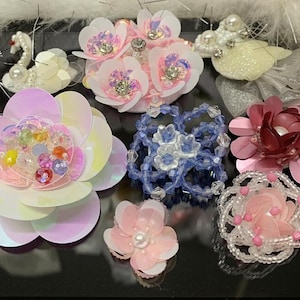 May include: Assortment of colorful fabric flower embellishments with sequins, beads, and pearls. The flowers are in various shades of pink, blue, white, and red.