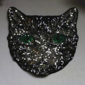 May include: A black cat face patch with green eyes made of sequins. The sequins are silver and gold.