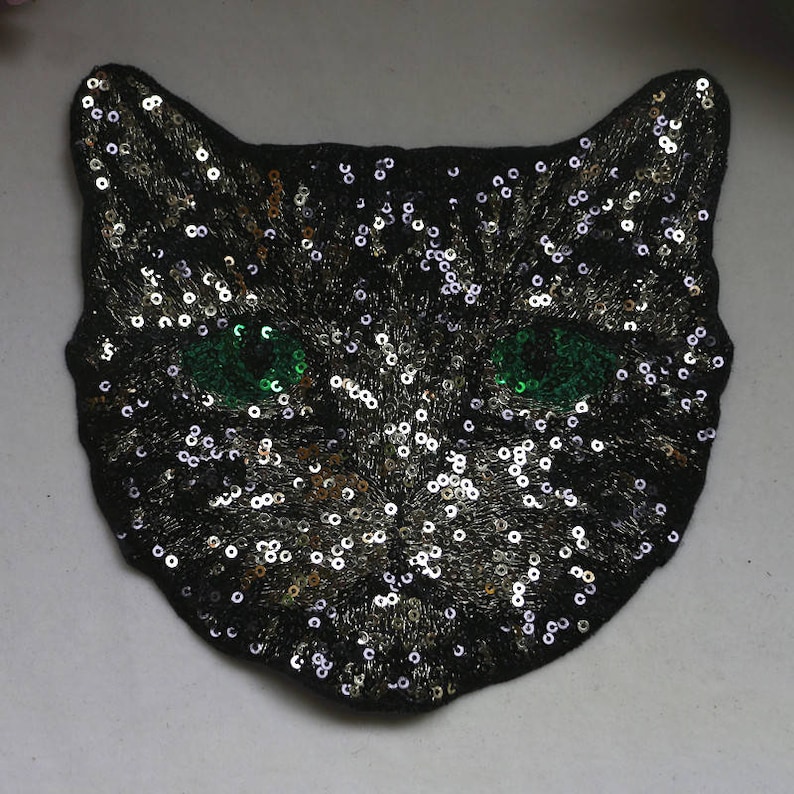 May include: A black cat face patch with green eyes made of sequins. The sequins are silver and gold.