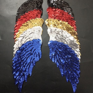 Peut inclure: Une paire d'ailes d'ange en sequins de six couleurs différentes : noir, rouge, or, argent, bleu et bleu foncé. Les ailes sont superposées, créant un effet multidimensionnel.