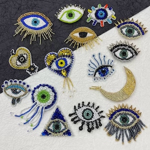 May include: A selection of beaded eye brooches in diverse designs. The brooches showcase a range of colours, including blue, green, gold, and white. Some have spike or fringe details, while others are heart-shaped or crescent-shaped.