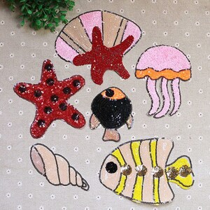 May include: A set of seven colorful iron-on patches featuring various sea creatures, including a starfish, a seashell, a jellyfish, a fish, and a clam. The patches are made of sequins and are perfect for adding a touch of whimsy to clothing, bags, or other accessories.