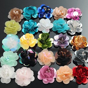 Puede incluir: Surtido de adornos de flores de tela de colores con lentejuelas y detalles de perlas. Las flores son de varios tonos de azul, verde, rosa, rojo, blanco y dorado.