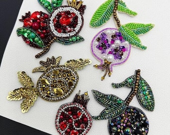 Puede incluir: Cinco broches decorativos con forma de granada elaborados con intrincados abalorios y lentejuelas. La colección presenta diseños en tonos rojos, morados, dorados e iridiscentes oscuros, cada uno con hojas de cuentas verdes y texturas.