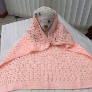 May include: A pink crocheted baby blanket with a textured pattern. The blanket is folded in half and a stuffed animal is peeking out from the fold.