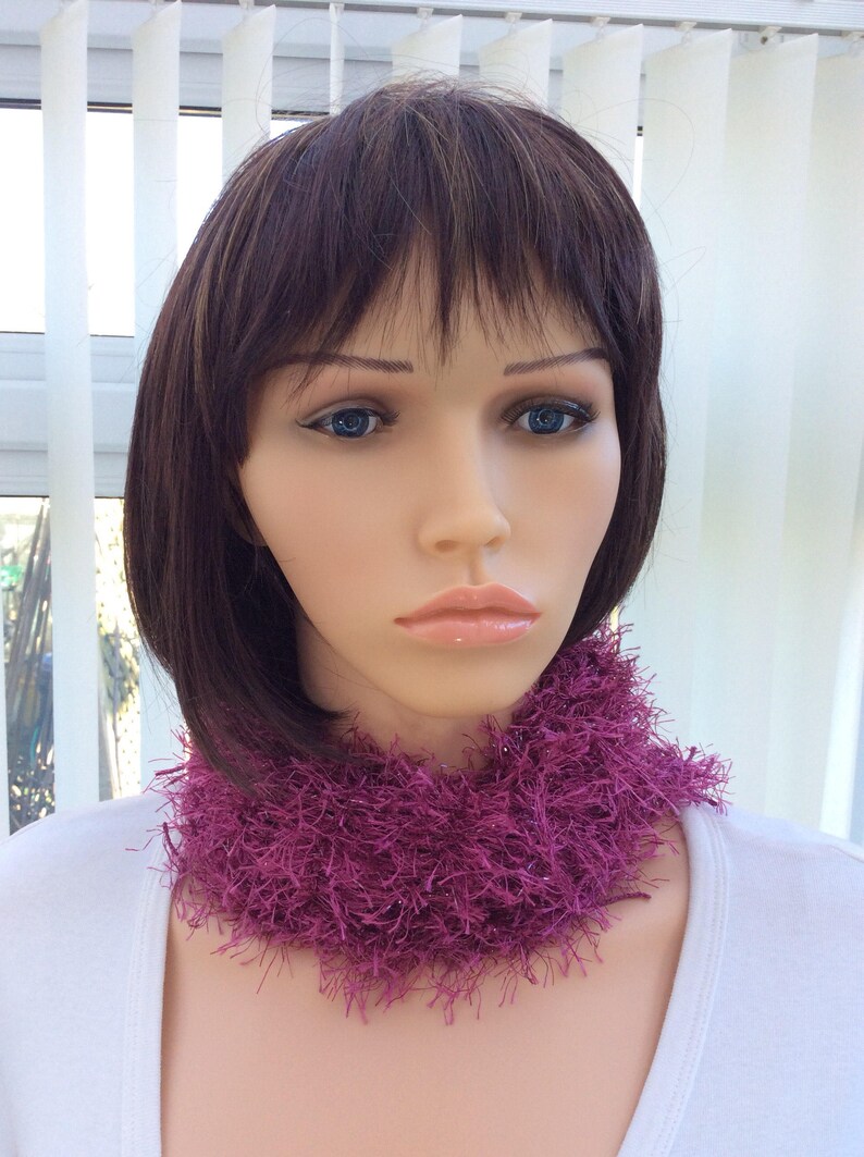 May include: A close-up of a mannequin wearing a pink fuzzy neck warmer. The neck warmer is made of a soft, fluffy material and has a loose, flowing design.