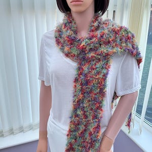 May include: A colorful, fuzzy scarf with a mix of red, orange, yellow, green, blue, and purple yarn. The scarf is long and thin, and it is draped around a mannequin's neck.
