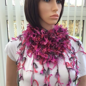 May include: A pink and grey fuzzy scarf with long fringe. The scarf is worn by a mannequin with short brown hair.
