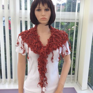 May include: A rust-colored crocheted shawl with fringe. The shawl is draped around a mannequin's neck.