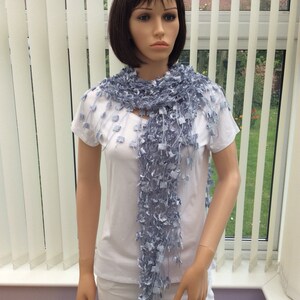 May include: A light blue crocheted scarf with a fringed design. The scarf is draped around the neck of a mannequin wearing a white t-shirt and white pants.
