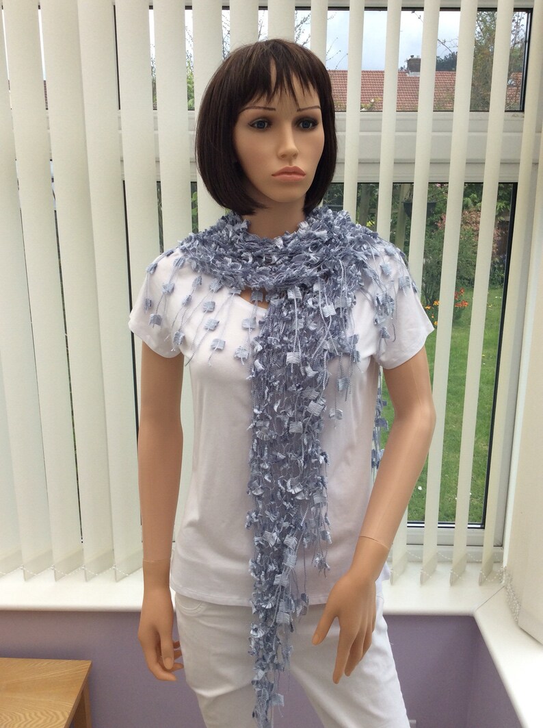 May include: A light blue crocheted scarf with a fringed design. The scarf is draped around the neck of a mannequin wearing a white t-shirt and white pants.