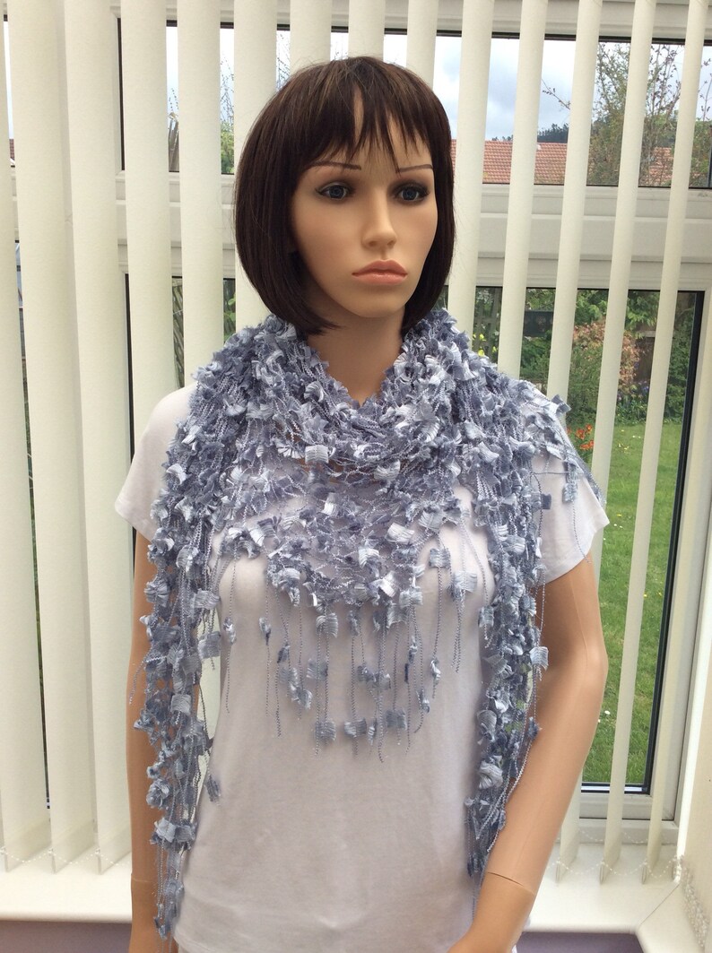May include: A light blue crocheted scarf with a delicate, lacy design and fringe. The scarf is draped around the neck of a mannequin.