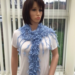 Peut inclure: Une &eacute;charpe en crochet bleu clair avec des d&eacute;tails en frange. L'&eacute;charpe est drap&eacute; autour du cou d'un mannequin.