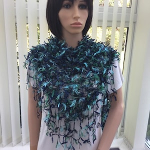 May include: A fringed shawl in teal, navy, and light blue. The handmade shawl is worn over a white top, highlighting its textured design. The fringe detail adds a delicate, flowing element to the accessory. The shawl is a stylish accessory.
