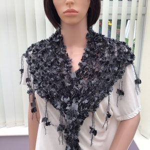 May include: A gray crocheted scarf with a fringed edge and a textured design. The scarf is draped around a mannequin's neck.