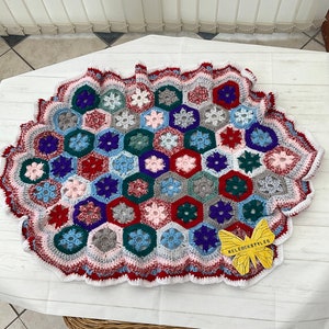 May include: A crocheted blanket with a hexagonal pattern in shades of red, blue, green, white, and gray. The blanket has a scalloped edge and is made with a variety of different stitches. The blanket is lying on a white surface with a small yellow butterfly sticker that says "KelebekStyles".