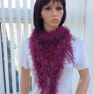 May include: A deep purple fuzzy scarf with long fringe. The scarf is worn by a mannequin with short brown hair.