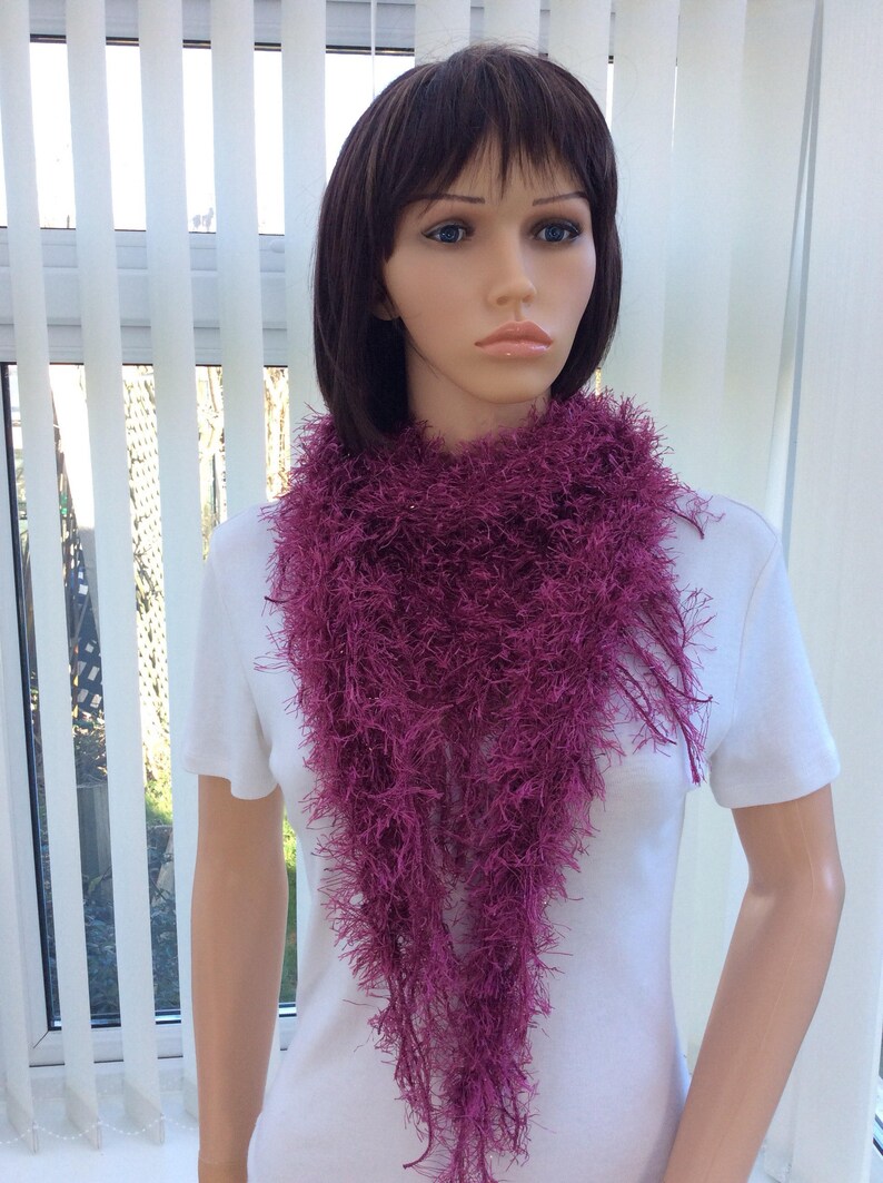 May include: A deep purple fuzzy scarf with long fringe. The scarf is worn by a mannequin with short brown hair.