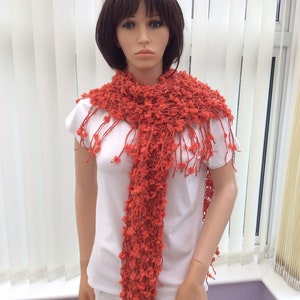May include: A coral colored knitted scarf with a fringe. The scarf is made of a soft, fluffy yarn and has a loose, open weave.