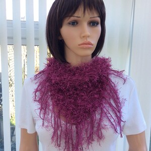 May include: A deep purple fuzzy scarf with long fringe. The scarf is worn by a mannequin with short brown hair.