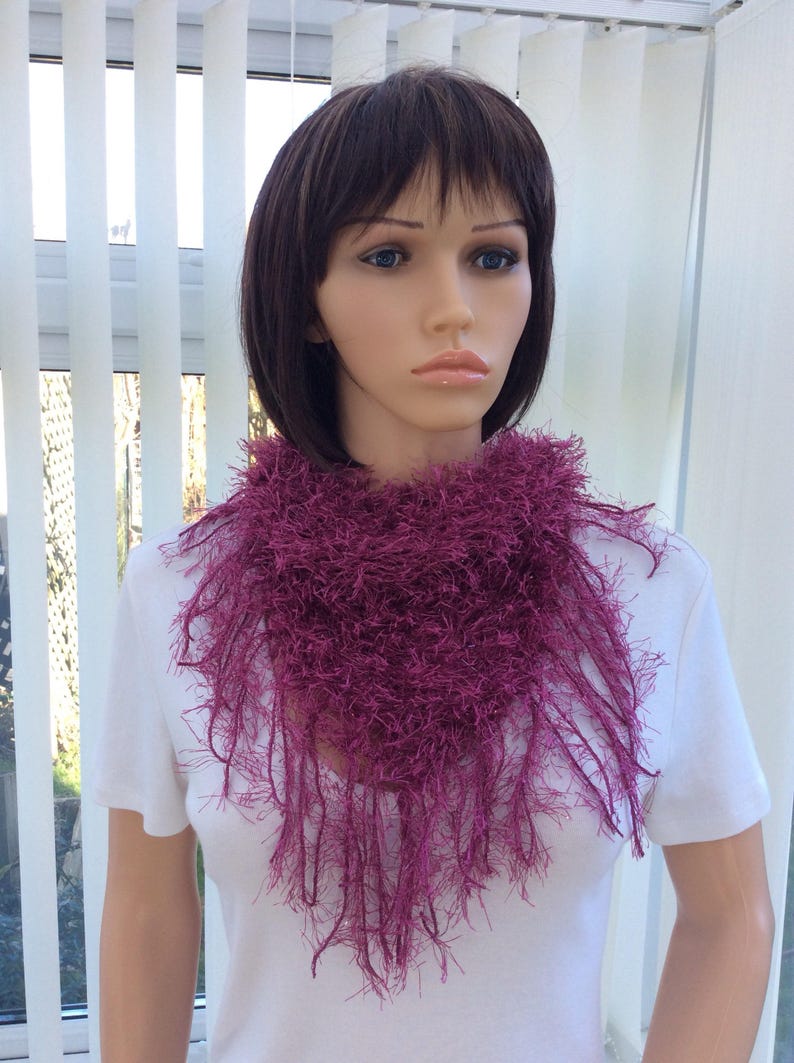 May include: A deep purple fuzzy scarf with long fringe. The scarf is worn by a mannequin with short brown hair.