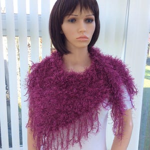 May include: A magenta fuzzy knit scarf with fringe. The scarf is draped around a mannequin's neck.