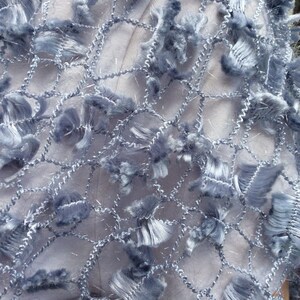 May include: A close-up of a light blue fabric with a net-like pattern and fringe details.