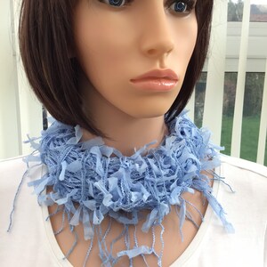 Peut inclure: Une &eacute;charpe frang&eacute;e bleu clair avec une texture tiss&eacute;e d&eacute;licate. L'&eacute;charpe est port&eacute;e autour du cou d'un mannequin aux cheveux courts et brun fonc&eacute;.