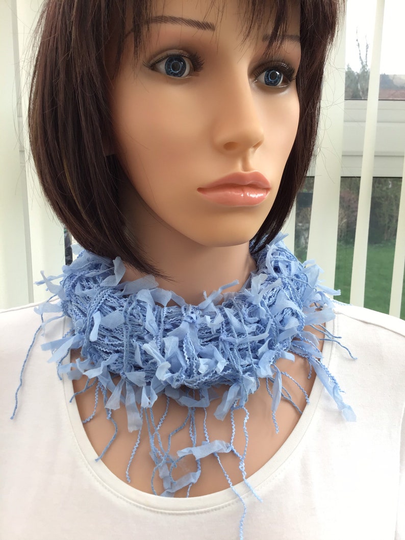 Peut inclure: Une &eacute;charpe frang&eacute;e bleu clair avec une texture tiss&eacute;e d&eacute;licate. L'&eacute;charpe est port&eacute;e autour du cou d'un mannequin aux cheveux courts et brun fonc&eacute;.