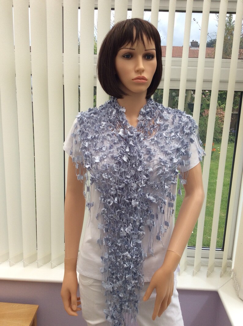 May include: A light blue crocheted shawl with a fringe. The shawl is draped over a mannequin's shoulders.