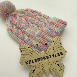 May include: A pastel pink, yellow, and blue knitted beanie with a pom pom on top. The beanie is pinned to a wooden butterfly with the text "KELEBEKSTYLES" written on it.