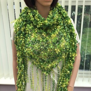 May include: A green and yellow scarf with a leaf-like pattern. The scarf is draped around a mannequin's neck and shoulders, with long fringe hanging down. The scarf's colours range from light yellow to dark green.