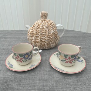 May include: A white teapot with a beige knitted tea cosy, accompanied by two matching teacups and saucers. The cups and saucers feature a floral design with pink edging. Small spoons are placed on the saucers. The set is on a grey tablecloth.