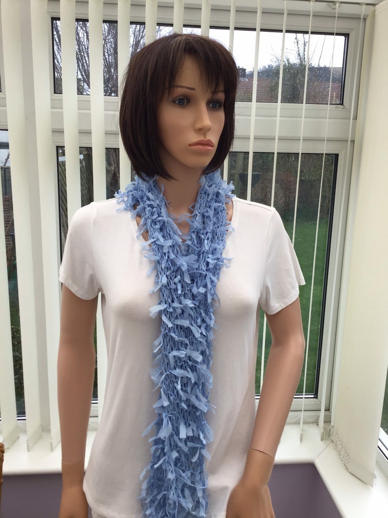 Peut inclure: Une &eacute;charpe bleu clair en tissu crochet&eacute; doux avec une bordure frang&eacute;e. L'&eacute;charpe est drap&eacute; autour du cou d'un mannequin.