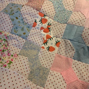 May include: A patchwork quilt featuring a variety of fabric patterns including small red strawberries on white, blue floral prints, pink gingham, and green leaf motifs. The textile is constructed from multiple geometric shapes stitched together.