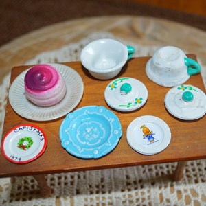 May include: Miniature wooden table set with a variety of small plates, cups, and lids. A pink and purple pastry sits on a white plate. Other plates are blue and white, some with floral designs. A small plate reads "CHRISTMAS 1979".