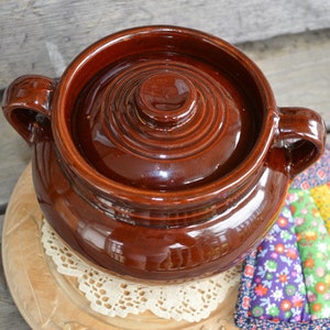 May include: A brown ceramic crock pot with a lid. The crock pot has two handles and is sitting on a wooden surface with a white doily.