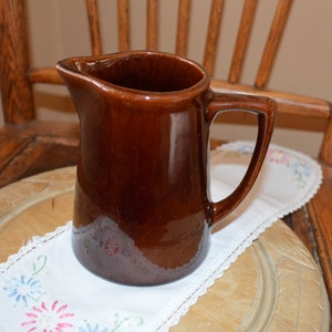 May include: A brown ceramic pitcher with a handle. The pitcher is sitting on a white doily with embroidered flowers.