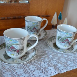 May include: Three white ceramic mugs with a winter scene design, each with a matching saucer. The mugs have a brown handle and are decorated with a red barn, a snowy bridge, and trees. The saucers are green and white with a floral pattern.
