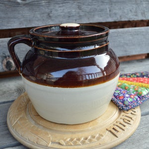 May include: A brown and white glazed ceramic bean pot with a lid. The pot is sitting on a wooden trivet with the word "Mama" carved into it.