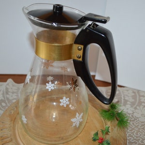 May include: A glass coffee pot with a gold band and a black plastic handle and lid. The pot is decorated with white snowflakes and has a clear glass body.