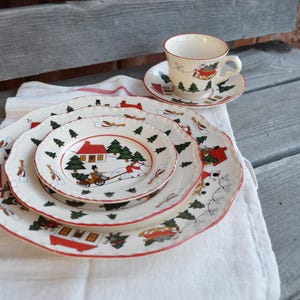May include: A vintage Christmas-themed dish set featuring a teacup and saucer, along with three plates of varying sizes. The white porcelain is decorated with red, green, and tan holiday scenes, including houses, trees, and figures.