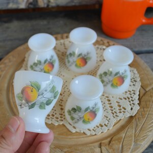 May include: Set of six white ceramic egg cups with a floral design featuring apples and leaves. The egg cups are arranged on a wooden tray with a lace doily.