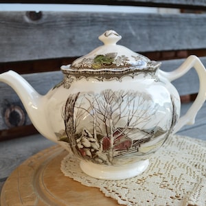 May include: Cream ceramic teapot with a winter scene illustration. The teapot has a lid with a floral design and a curved handle. The illustration depicts a snowy landscape with a red barn.