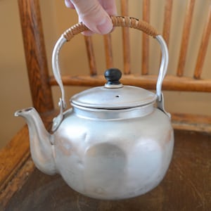 May include: A silver aluminum kettle with a brown woven handle and a black knob on the lid. The kettle has a slightly rounded, octagonal shape.