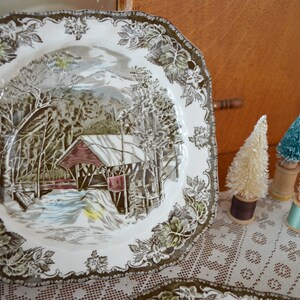 May include: A square, white ceramic plate with a brown and green floral border. The center of the plate features a winter scene with a covered bridge, trees, and a river.